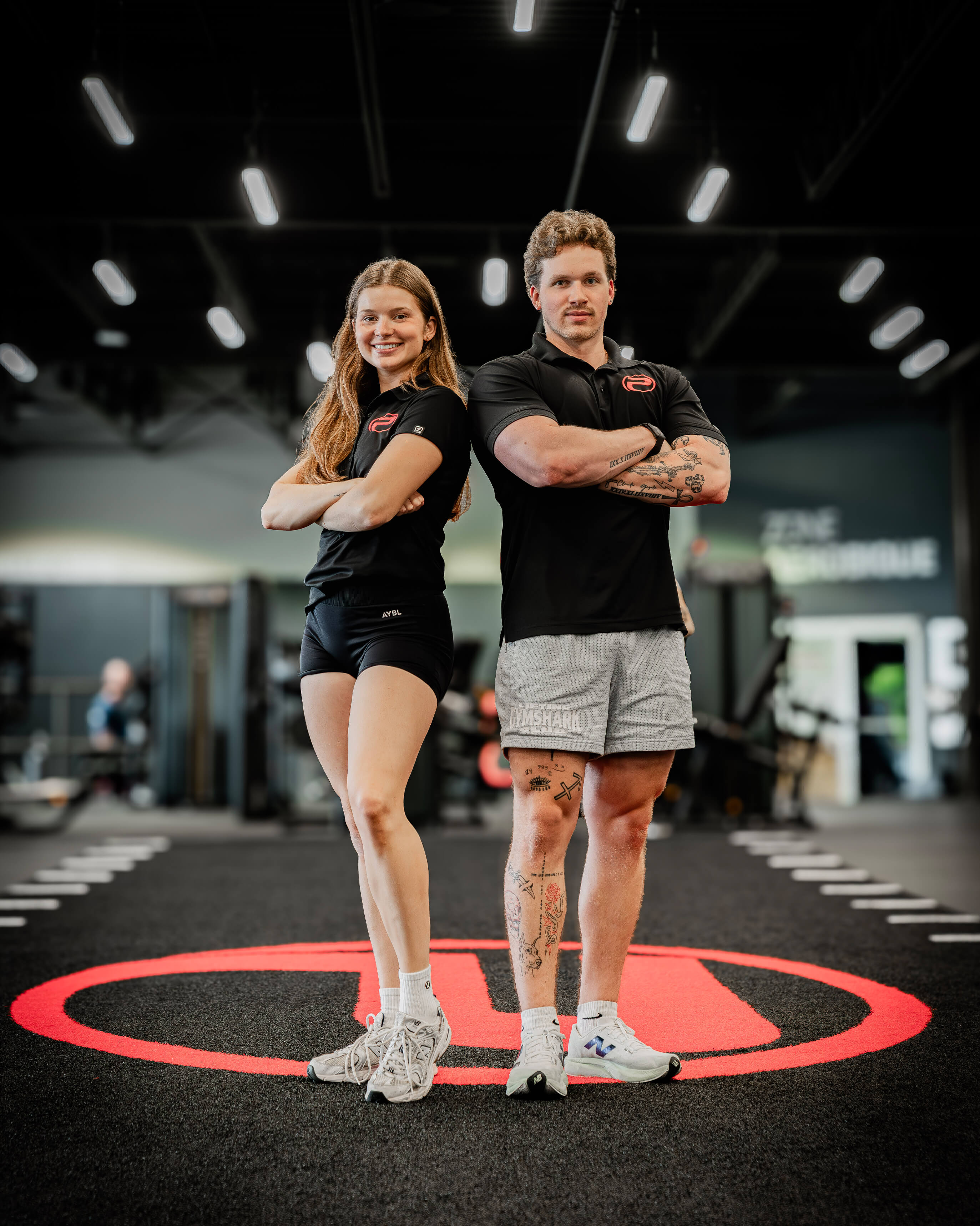 Un homme et une femme, posent côte à côte, bras croisés, portant un polo ProGym, sur un tapis noir avec le grand logo rouge du ProGym au sol, à l’intérieur d’une salle d’entraînement moderne.